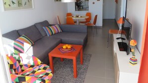 Een moderne woonkamer met een grijze modulaire bank, kleurrijke kussens, een klein oranje tafeltje met fruit, en een levendige fauteuil met een geometrisch patroon.