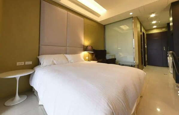A modern hotel room with a large bed, a bedside table, a lamp, and a glass partition wall.