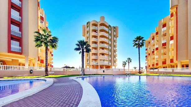 A swimming pool with a brick walkway, surrounded by palm trees and modern apartment buildings. - 6th floor apartment ideal for families, free wifi, balcony, sea views (La Manga del Mar Menor)