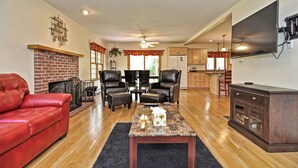 A spacious living room with a red leather sofa, a black leather chair, and a coffee table. There is a fireplace, a television, and a kitchen area in the background. - Little Sandy Pond - Plymouth Estate • Family & Exec Stays • Sleeps 16 (Plymouth)