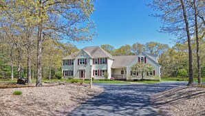 A two-story house with a front porch, surrounded by trees and a gravel driveway. - Little Sandy Pond - Plymouth Estate • Family & Exec Stays • Sleeps 16 (Plymouth)