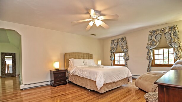 A bedroom with a large bed, a nightstand, a ceiling fan, and a window with curtains. - Little Sandy Pond - Plymouth Estate • Family & Exec Stays • Sleeps 16 (Plymouth)