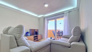 A living room with two grey sofas, a wooden TV stand, and a balcony with a table and chairs. - Family 5th floor apartment, sea views, balcony, communal pool, padel (La Manga del Mar Menor)