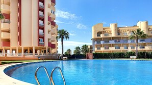 A swimming pool with a clear blue water surface, surrounded by a brick walkway. In the background, there are multi-story buildings with balconies and a palm tree. - Family 5th floor apartment, sea views, balcony, communal pool, padel (La Manga del Mar Menor)