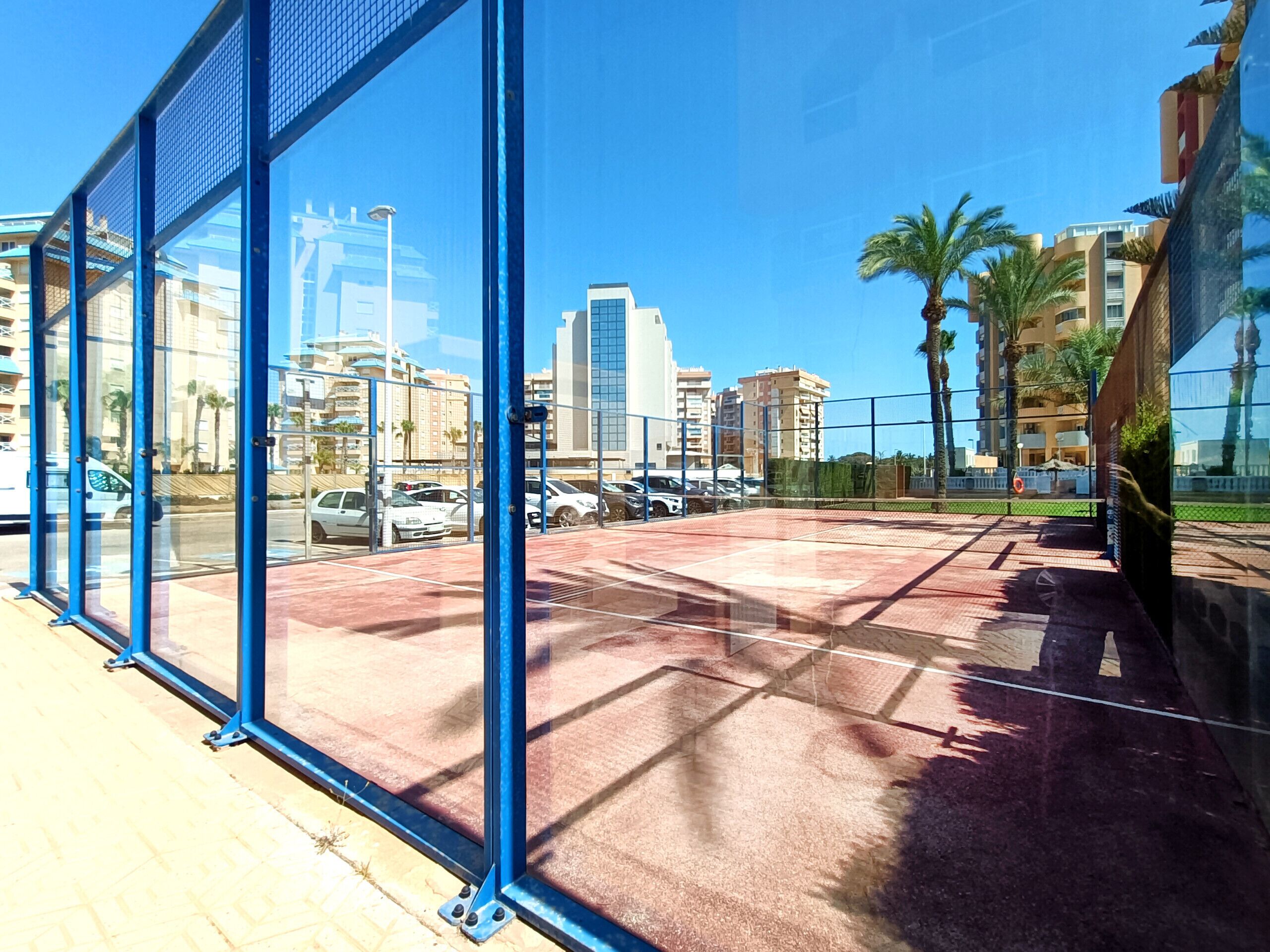 A view through a glass door of a parking area with a red pavement, surrounded by buildings and palm trees.