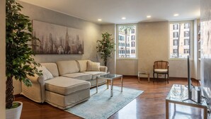 Ein Wohnzimmer mit einer Couch, einem Couchtisch, einem Sessel, einem Fernseher und einem großen Fenster mit Blick auf Gebäude.