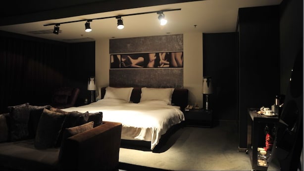 Ein modernes Hotelzimmer mit einem großen Bett, einer Sitzecke mit einer Couch und Sesseln, einem Nachttisch mit Lampe sowie einem an der Wand befestigten Kunstwerk.