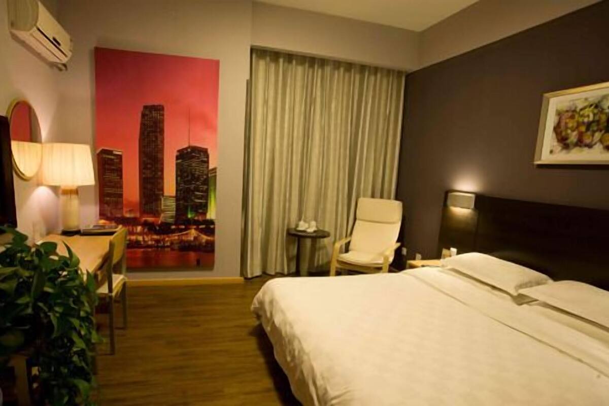 Habitación de hotel con cama, escritorio con lámpara, silla, mesita y planta en maceta.