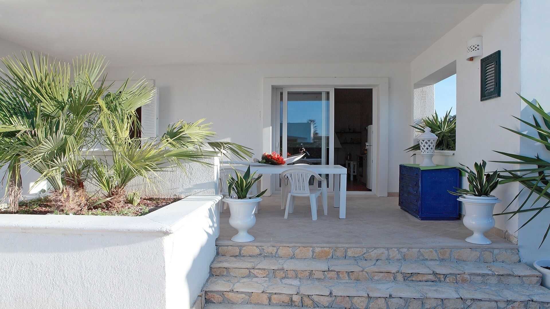 Fachada blanca con una escalera de piedra que lleva a una puerta, mobiliario exterior blanco y plantas en macetas.