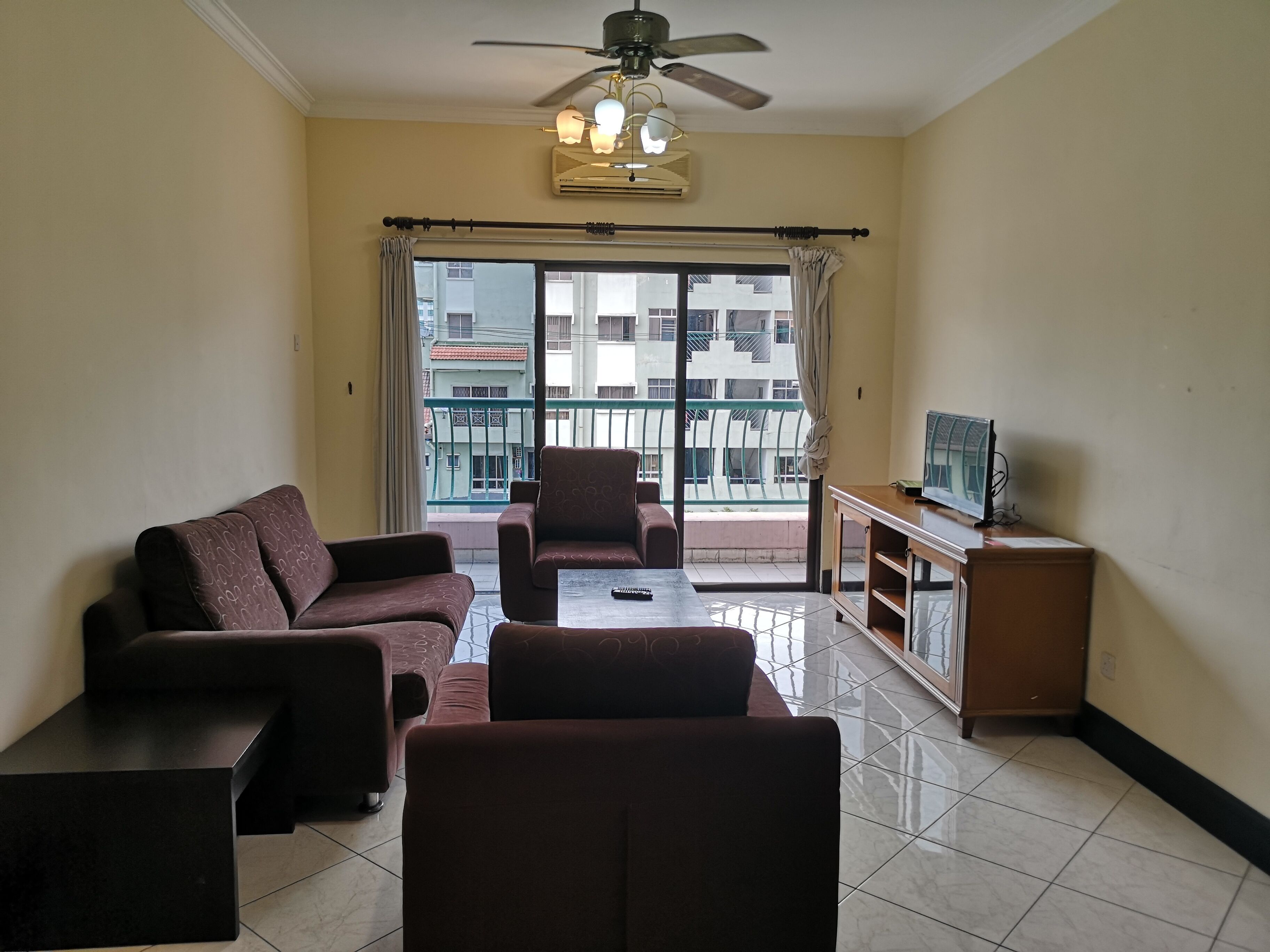 A living room with a sofa set, a coffee table, a TV on a stand, and a balcony with a view of buildings.