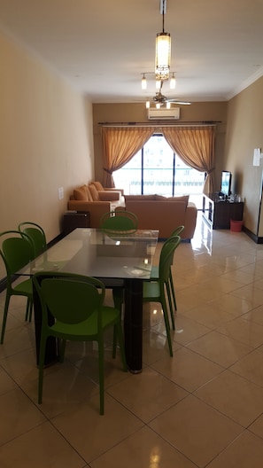 A spacious living room with a large glass table surrounded by green chairs, a sofa, and a ceiling fan.