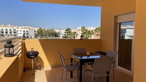 A balcony with a table and chairs, a grill, and a view of a residential area with palm trees and buildings.
