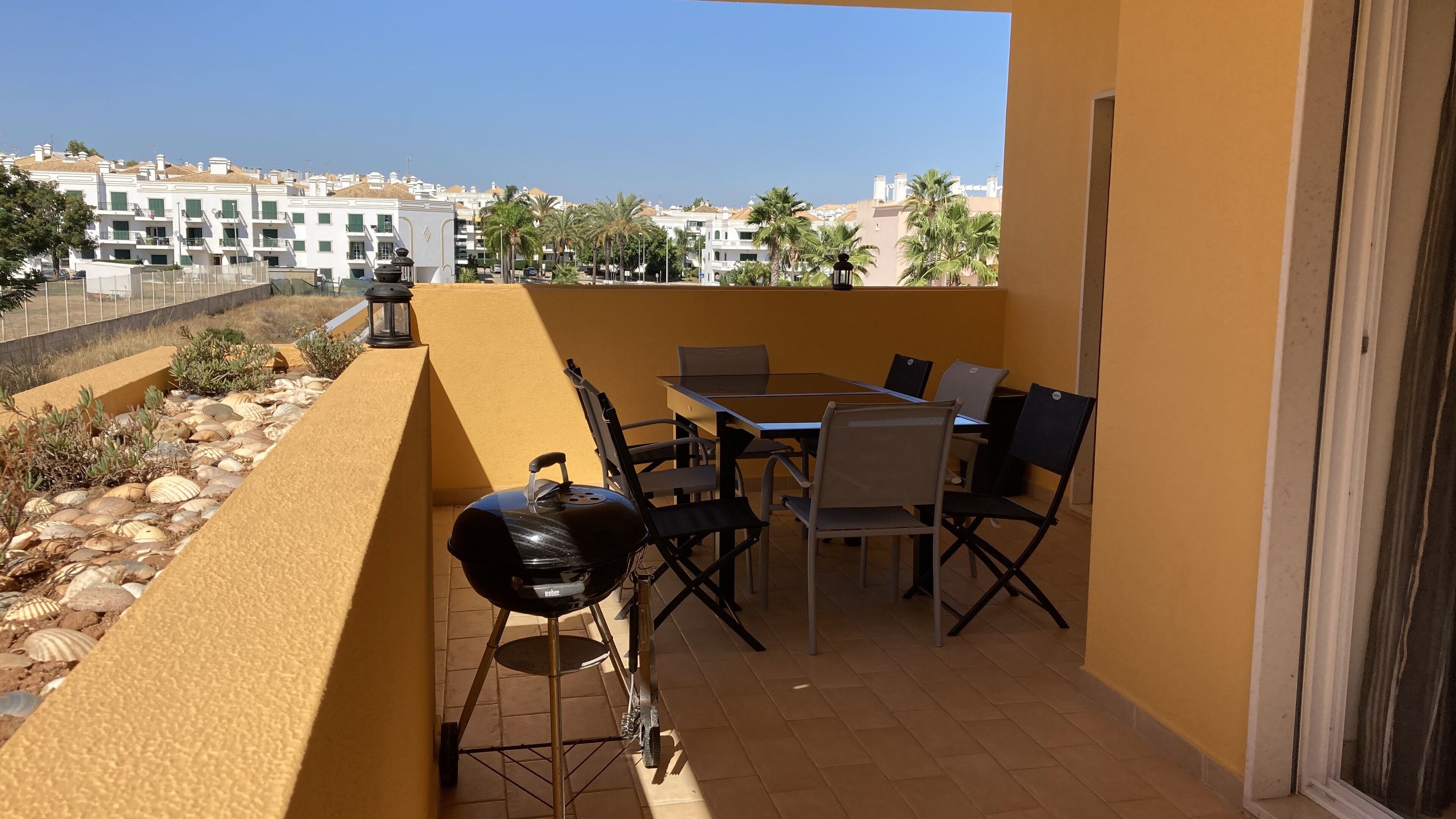 En balkon med et bord, stole, grill og udsigt til bygninger og palmer.