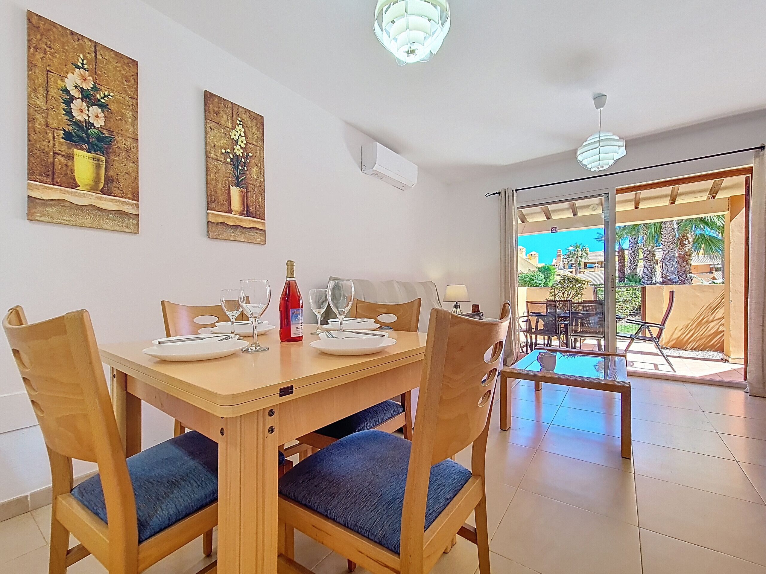 A dining area with a wooden table set for four, chairs with blue cushions, two wall-mounted paintings, and a view of an outdoor patio with tables and chairs.