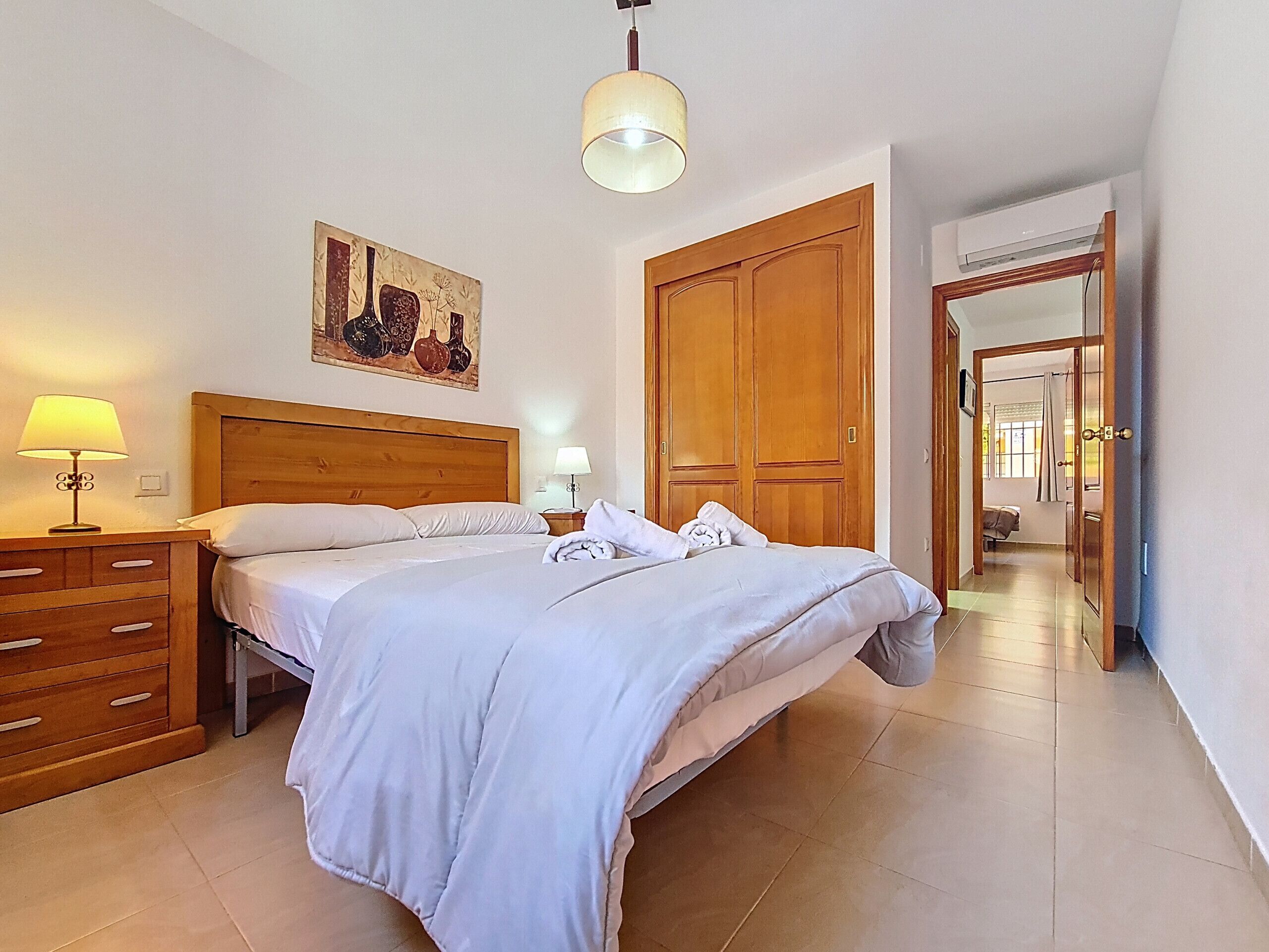A hotel room with a bed, wooden headboard, bedside lamps, a painting, a wardrobe, and a bathroom visible through an open door.