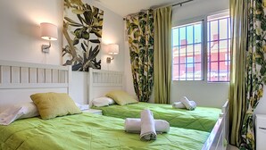 Habitación de hotel con dos camas, ropa de cama verde, cabecera blanca, lámparas fijadas a la pared y una ventana con cortinas.