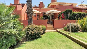Una casa de fachada roja, con un porche cubierto y sombrillas, y un jardín cuidado con diversas plantas y flores.