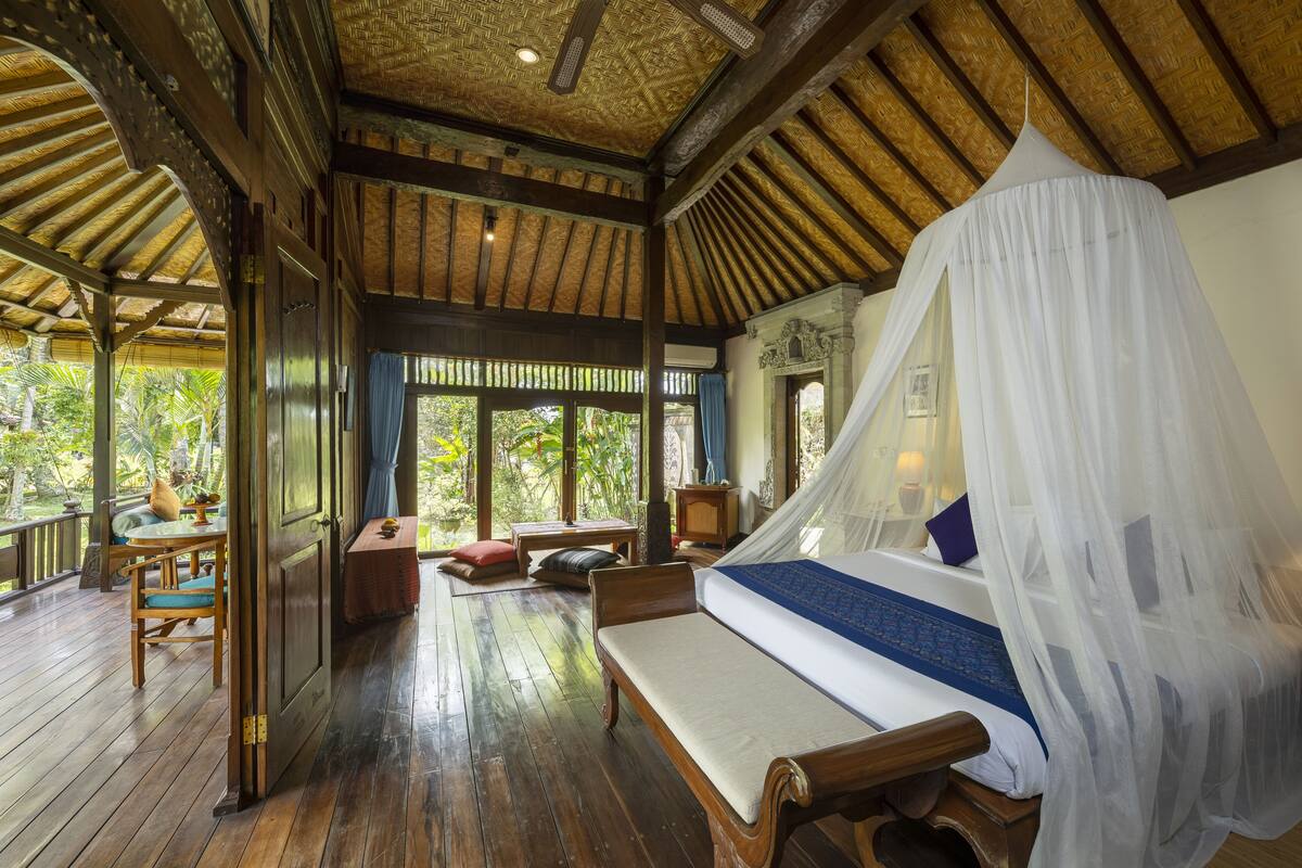 A room with a bed under a canopy, wooden flooring, a bench, a small table, and a view of greenery outside.