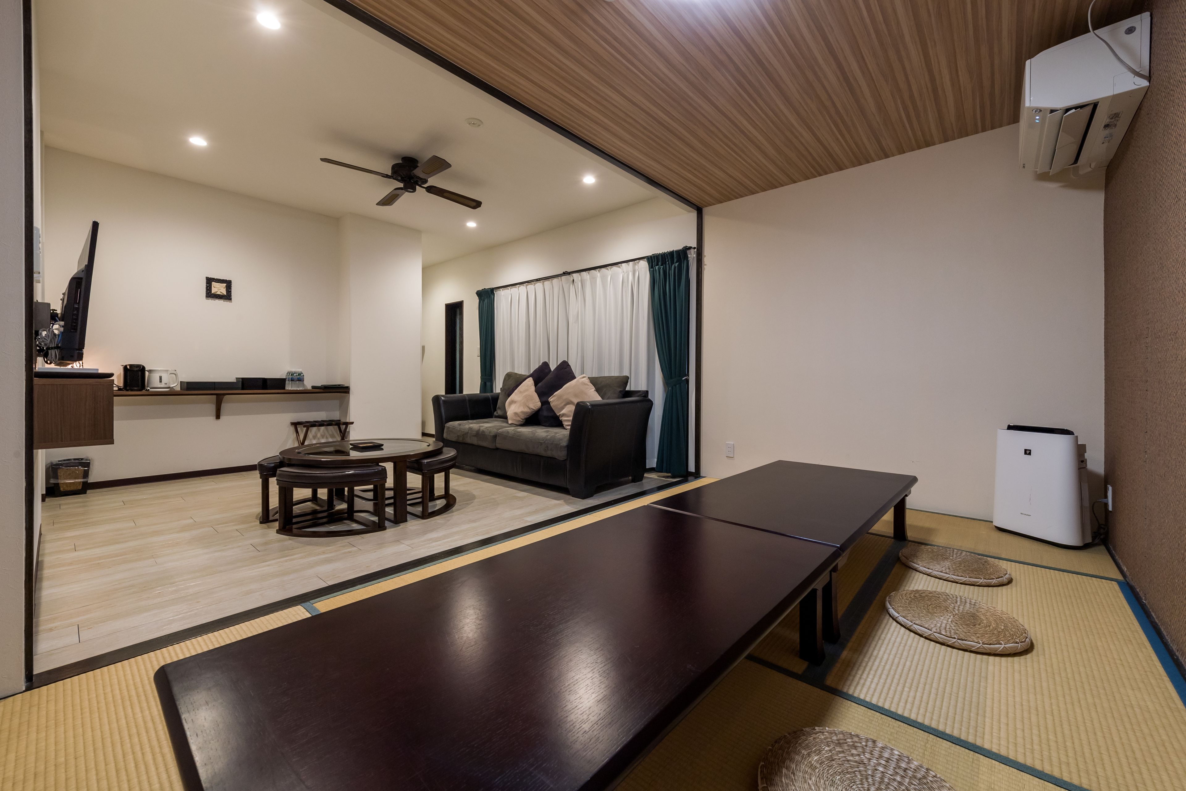 A modern living room with a long dark wooden table, a sofa with cushions, a ceiling fan, and a kitchen area in the background.