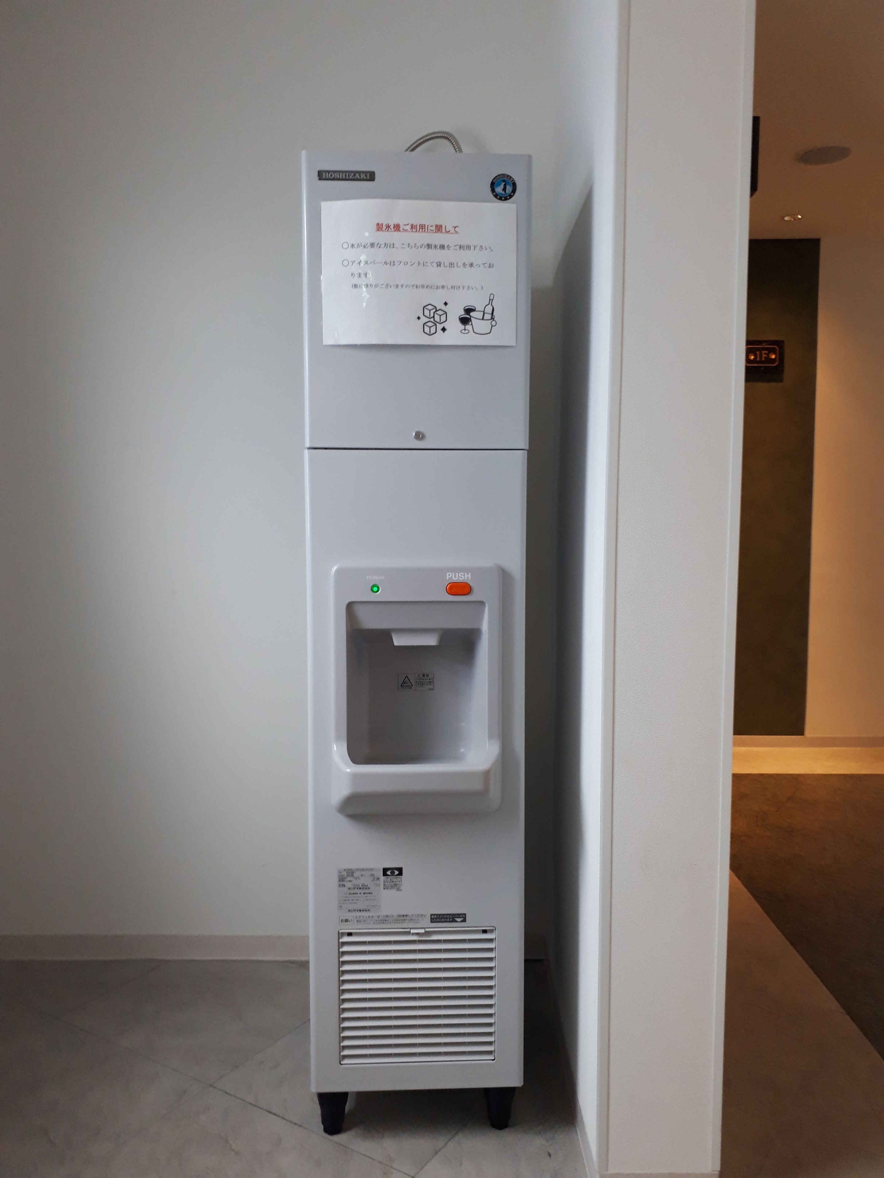 A white vending machine with a water dispenser, located in a hallway with a tiled floor.