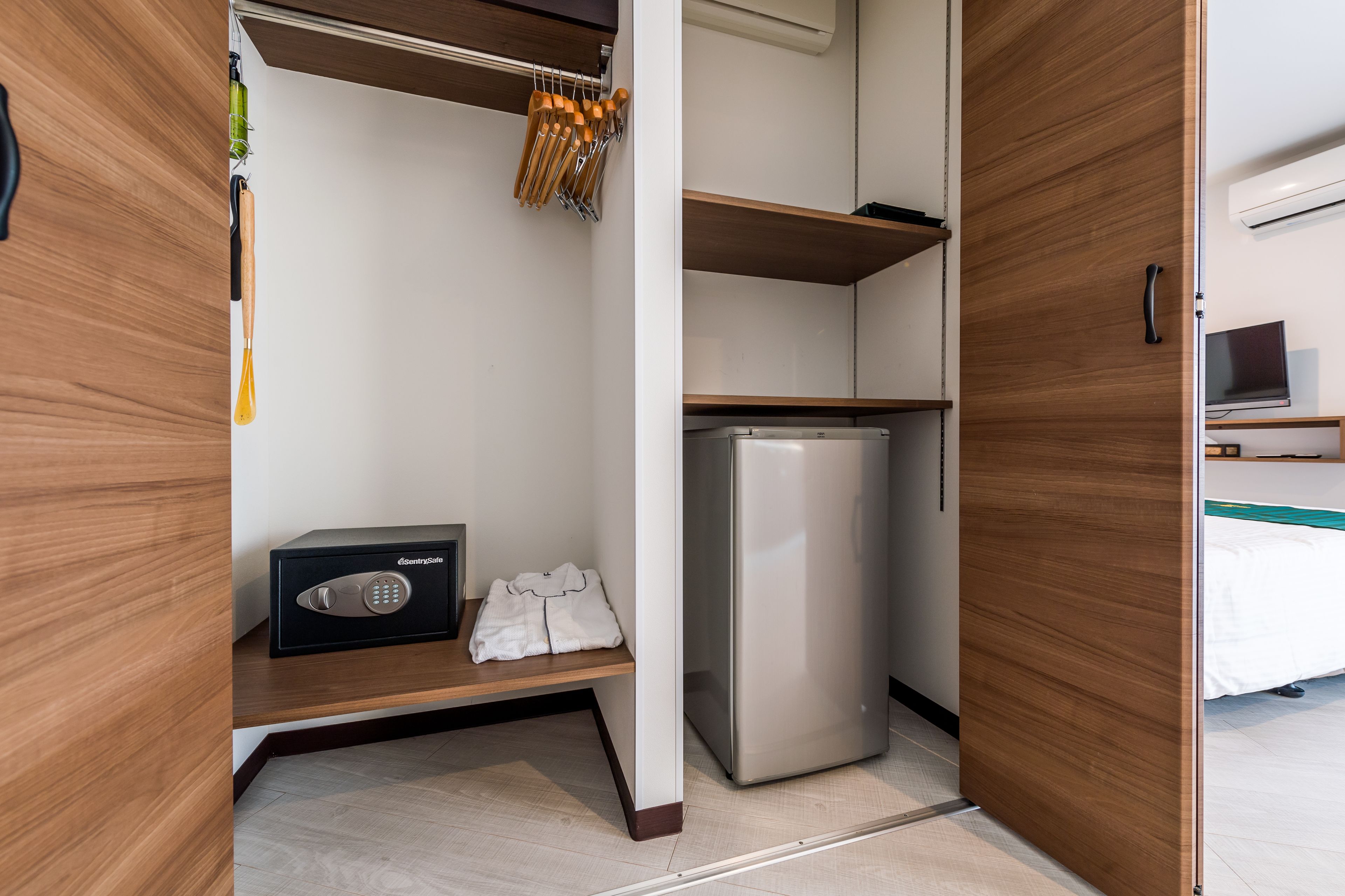 A hotel room with a wooden wardrobe, a small refrigerator, a black safe, and a shelf with a folded white towel.