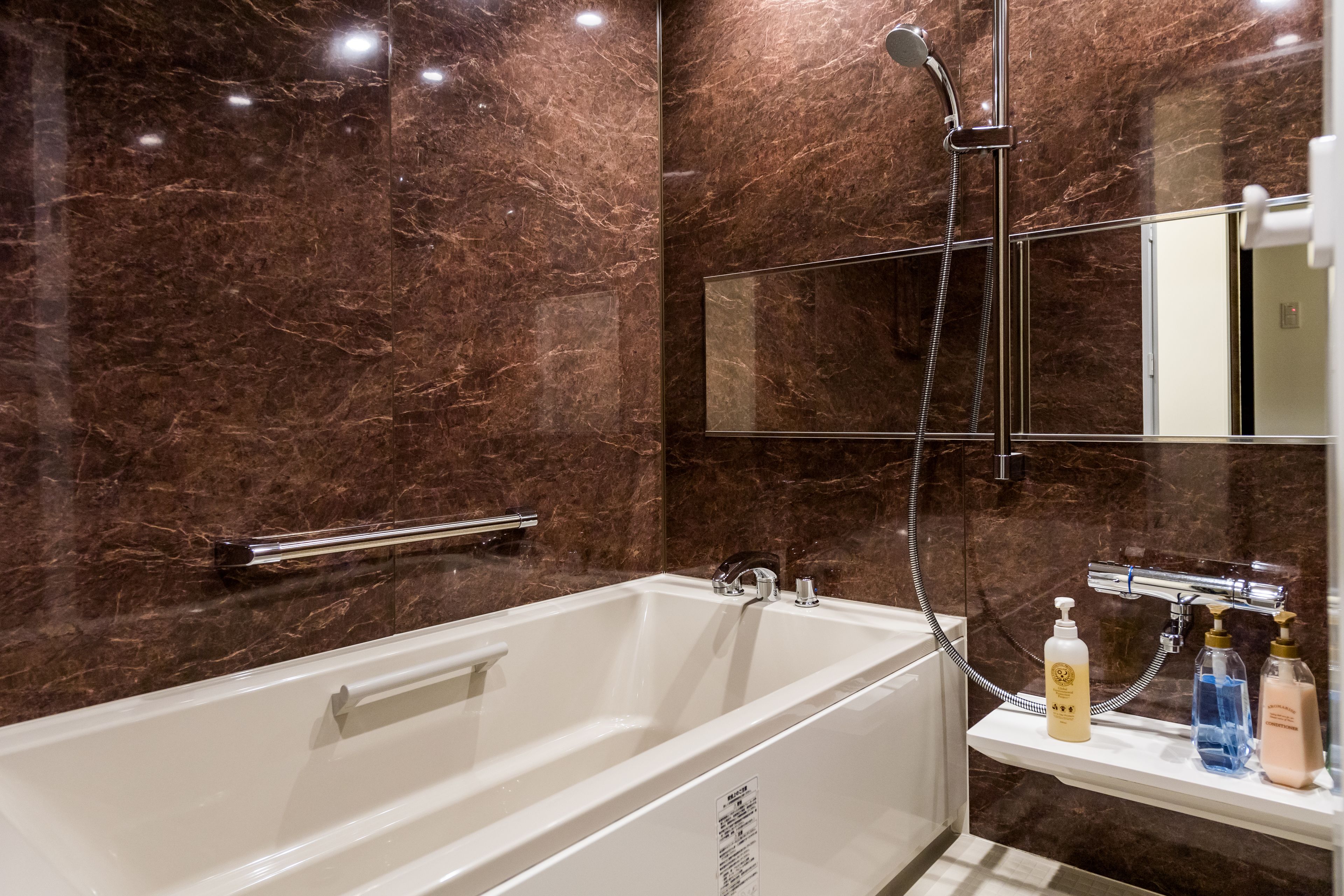 A modern bathroom with a marble wall, a white bathtub, a handheld showerhead, and a shelf with toiletries.