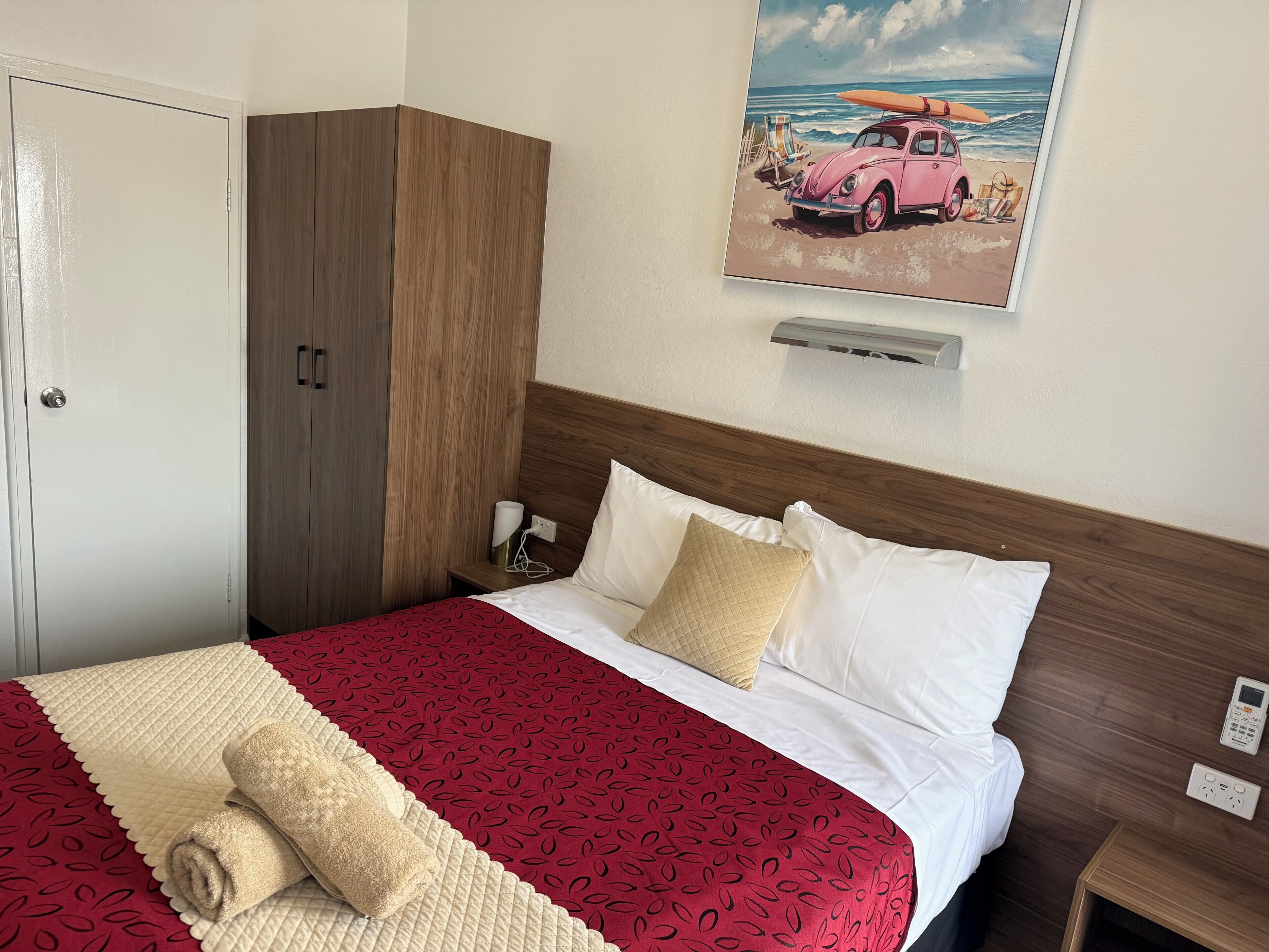 A hotel room with a bed, a wooden wardrobe, a nightstand, a wall-mounted air conditioner, and a beach-themed painting.