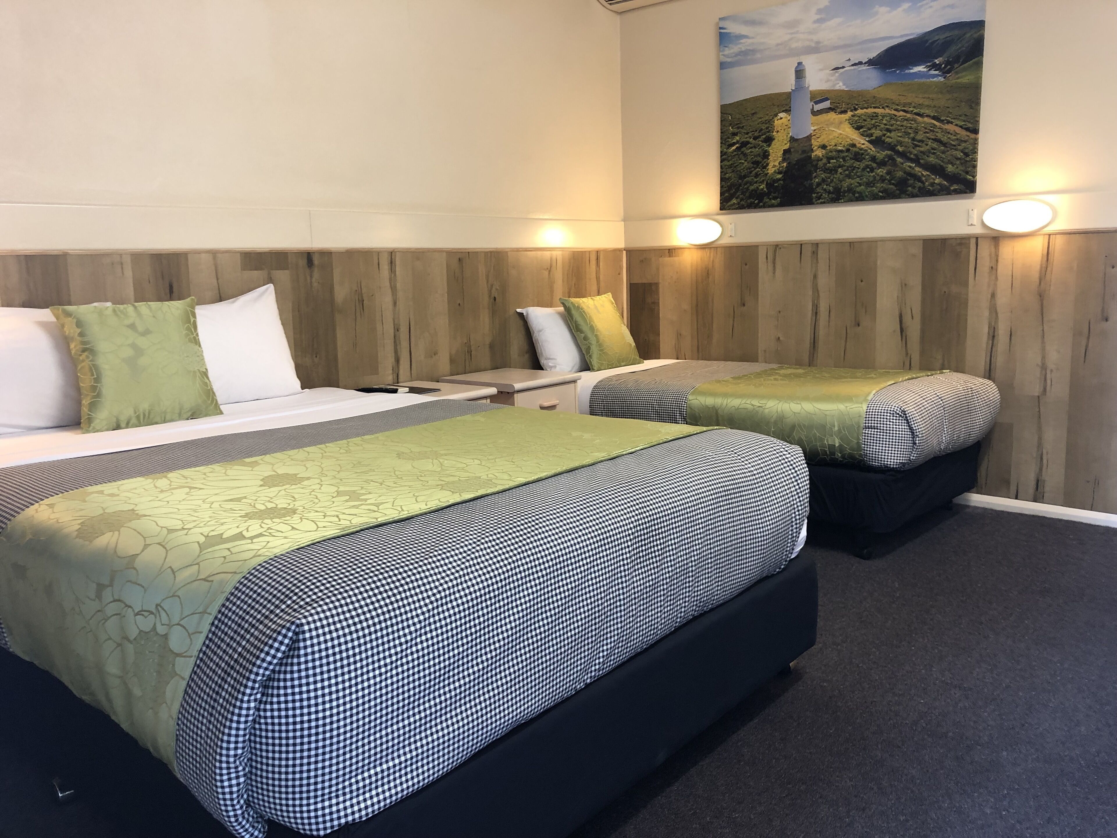 Quarto de hotel com duas camas, cada uma com colcha xadrez e travesseiro verde. Há uma pintura de um farol na parede acima das camas.