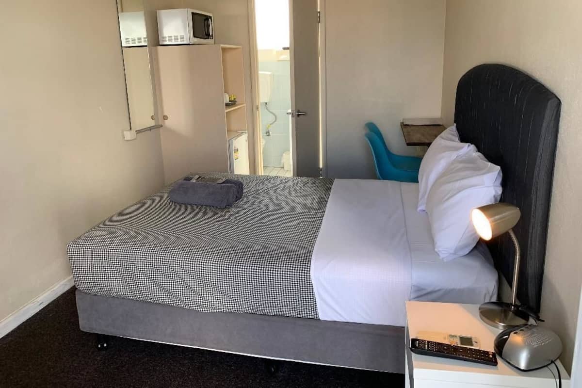 A single bed with a checkered bedspread, a bedside lamp, a remote control, and a small table with a book and a cup.