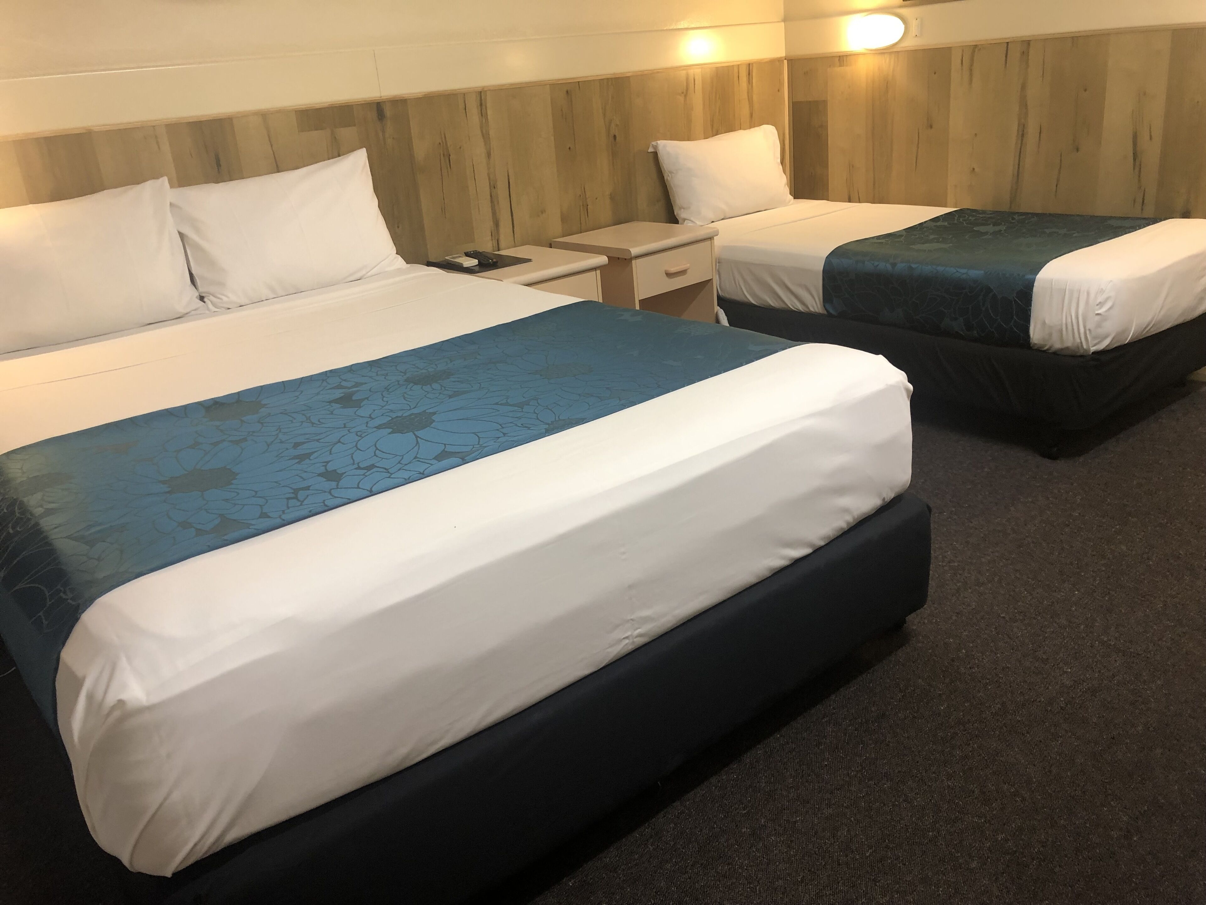 A hotel room with two beds, each with a white and blue bedspread, a wooden headboard, a nightstand, and a small table.