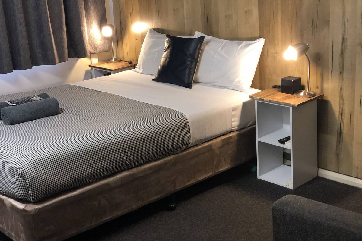 A modern hotel room with a wooden headboard, a large bed with a checkered bedspread, two bedside lamps, and a small bedside table with a lamp.