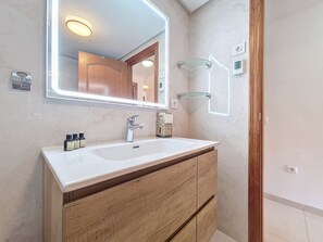 Ein Badezimmer mit einem weißen Waschbecken, einer Holzwaschmöbel-Anrichte und einem Spiegel mit Beleuchtung. Über dem Waschbecken befinden sich zwei Glasregale und rechts eine Dusche.
