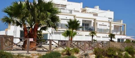 A modern multi-story building with balconies, surrounded by palm trees and a sandy landscape.