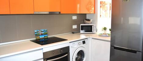 A compact kitchen with white and orange cabinets, a stainless steel refrigerator, a microwave, a washing machine, and a dishwasher.
