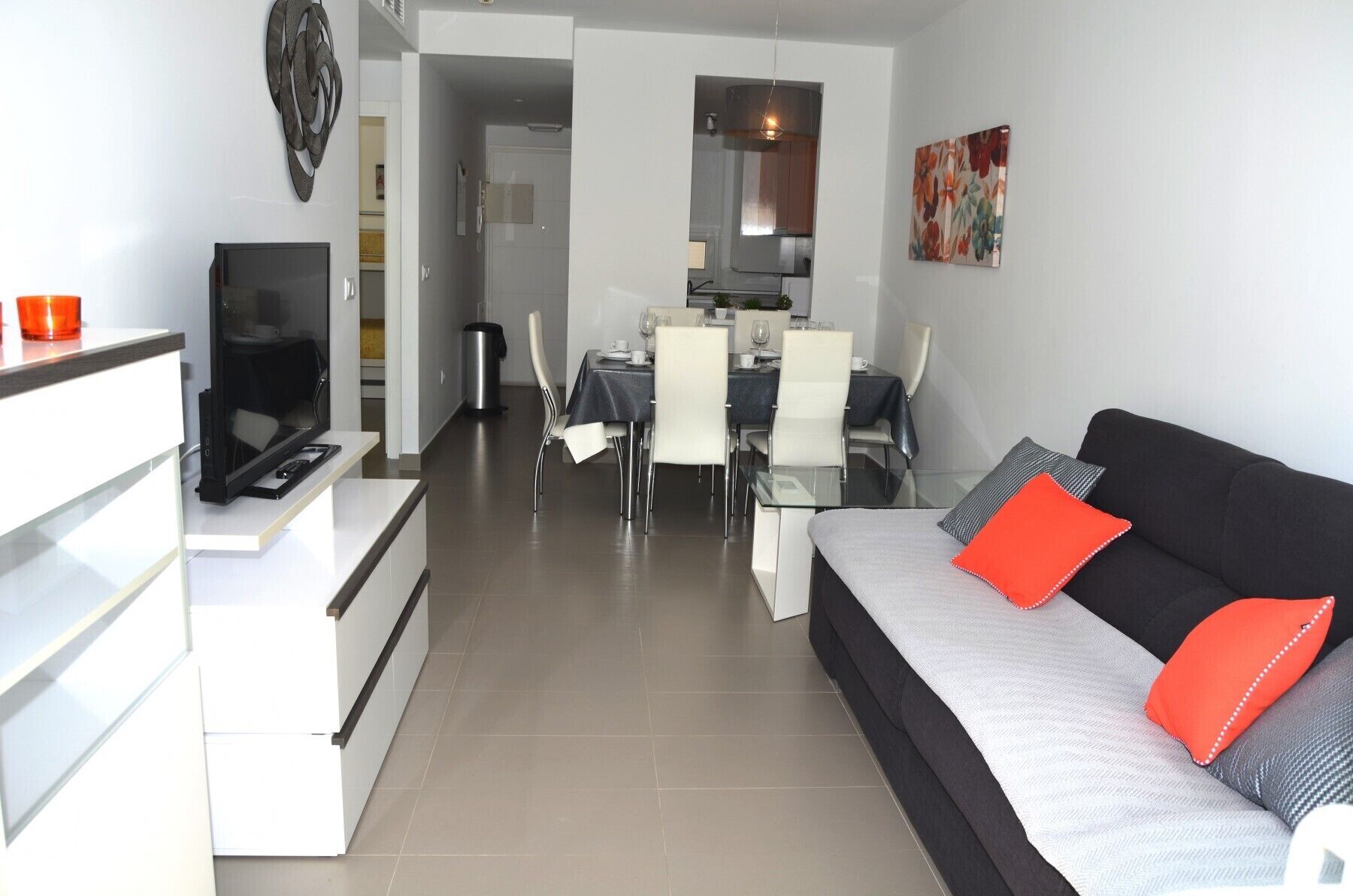 A modern living room with a black sofa, white and grey cushions, a dining area with a table and chairs, and a kitchen in the background.