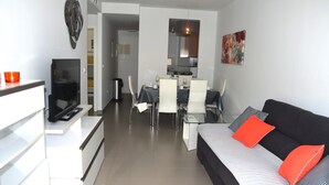 A modern living room with a black sofa, white and grey cushions, a dining area with a table and chairs, and a kitchen in the background. - Ground floor apartment, patio, free wifi, sea view, BBQ (La Manga del Mar Menor)