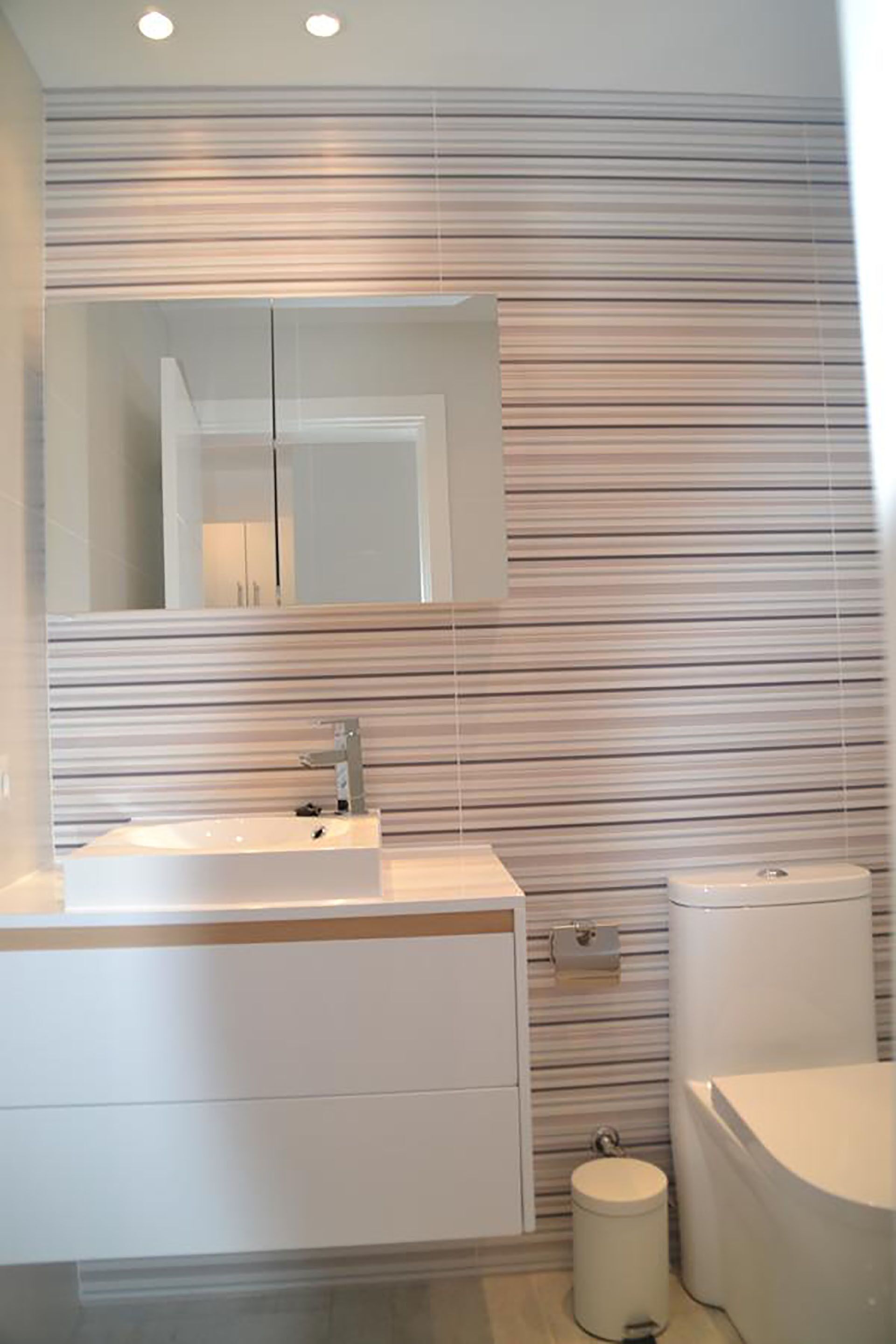 Een moderne badkamer met een gestreept wandontwerp, een grote spiegel, een wit wastafelmeubel en een toilet.