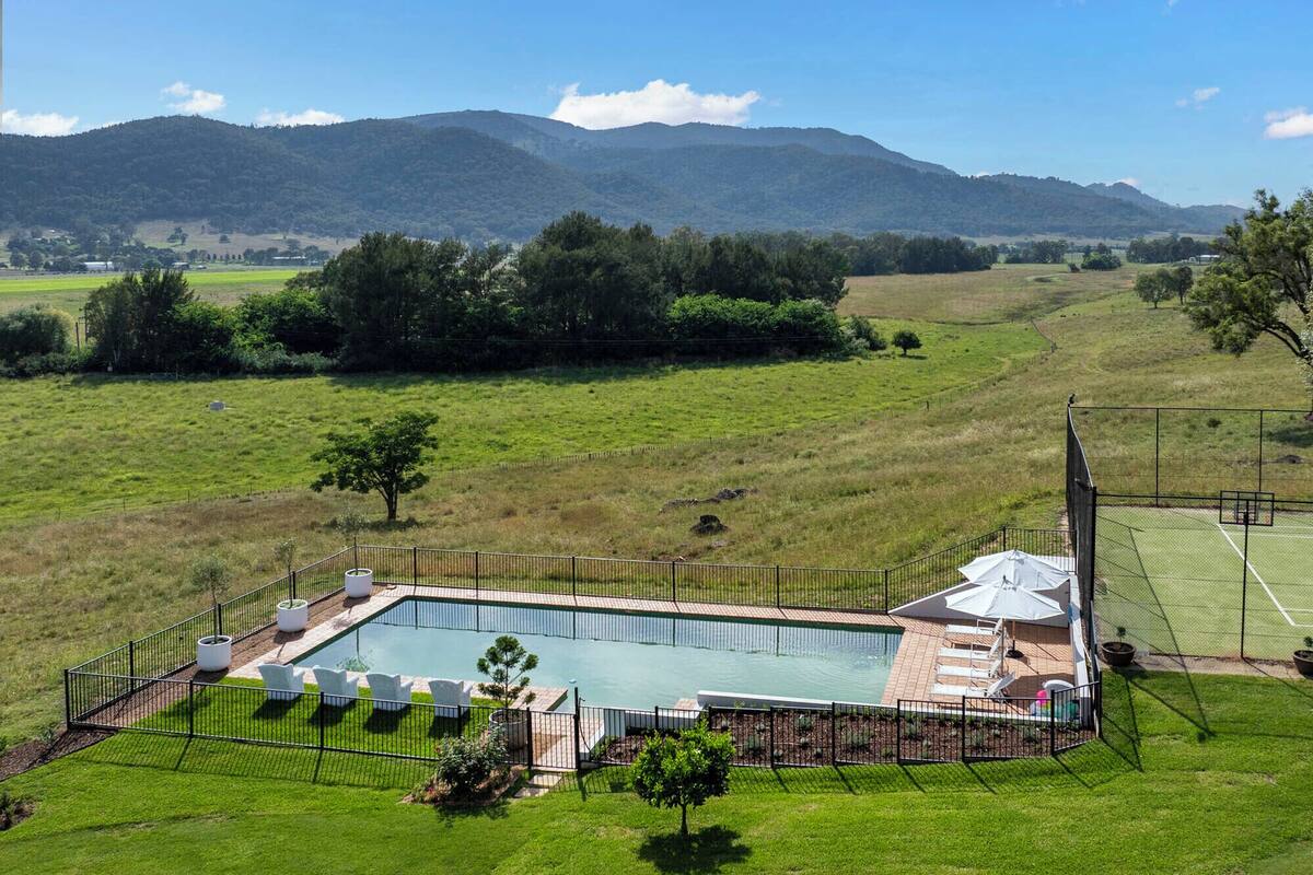 Une piscine rectangulaire dotée d’un parasol et de chaises longues, entourée d’une clôture, avec en arrière-plan de vastes champs verdoyants et des montagnes.