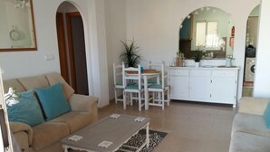 Un salon avec un canapé beige, une table basse blanche et une table à manger avec des chaises. Il y a également un coin cuisine avec des meubles blancs et un évier.