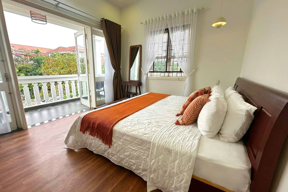 Ein Schlafzimmer mit Bett, einem Balkon, einem Fenster mit Vorhängen und Blick ins Grüne.