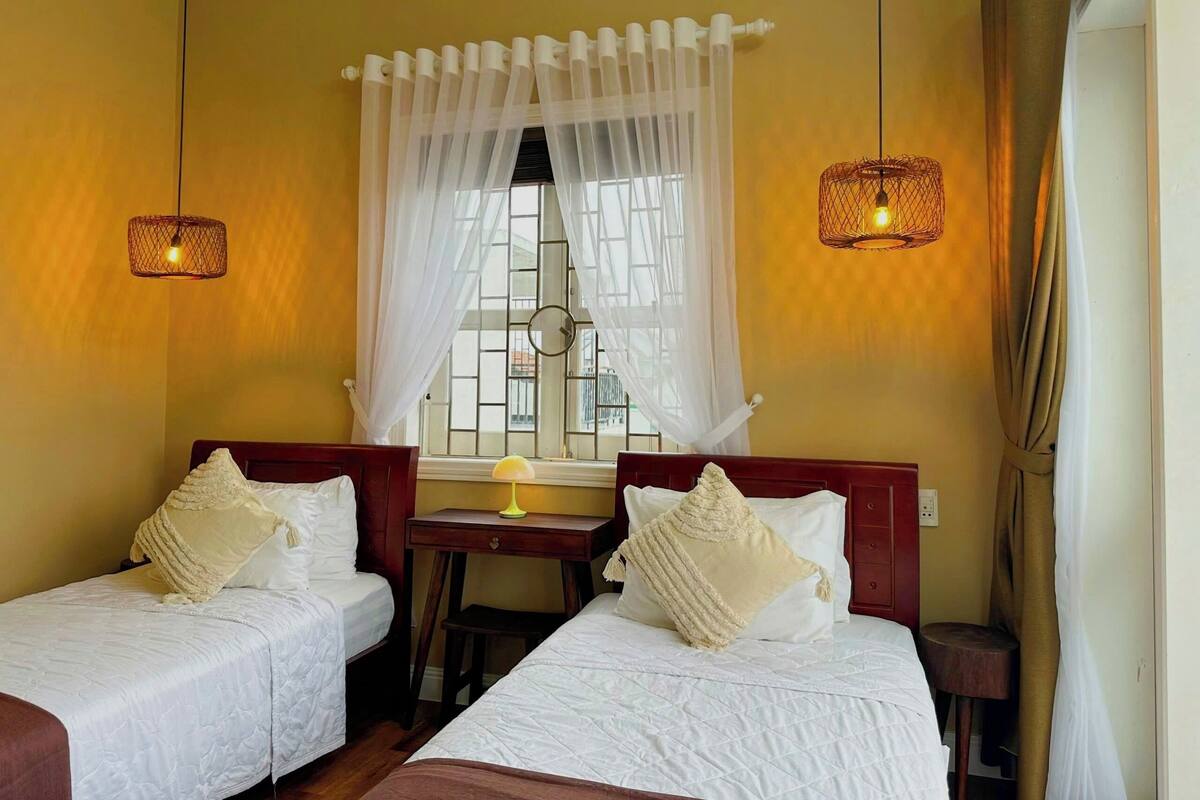 Ein Hotelzimmer mit zwei Einzelbetten, einem kleinen Schreibtisch mit Lampe, einem Fenster mit weißen Vorhängen und zwei Wandleuchten.