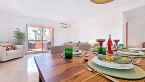 Un coin repas avec une table en bois dressée pour le repas, un canapé et une vue sur les palmiers, visible à travers une porte coulissante.