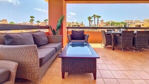 Une terrasse sur le toit, aménagée avec des meubles en osier, une table à plateau de verre, et offrant une vue sur les palmiers et les immeubles.