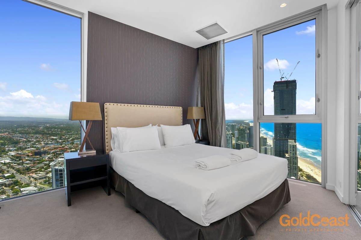 A spacious bedroom with a large bed, two bedside tables, and a view of a cityscape and beach.