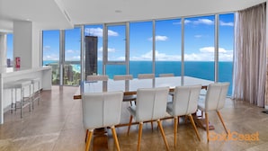 A modern dining area with a large table and chairs, large windows with a view of the ocean and cityscape.
