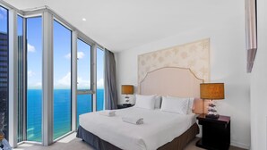 A hotel room with a large bed, two bedside tables, a headboard with a floral pattern, and a view of the ocean.