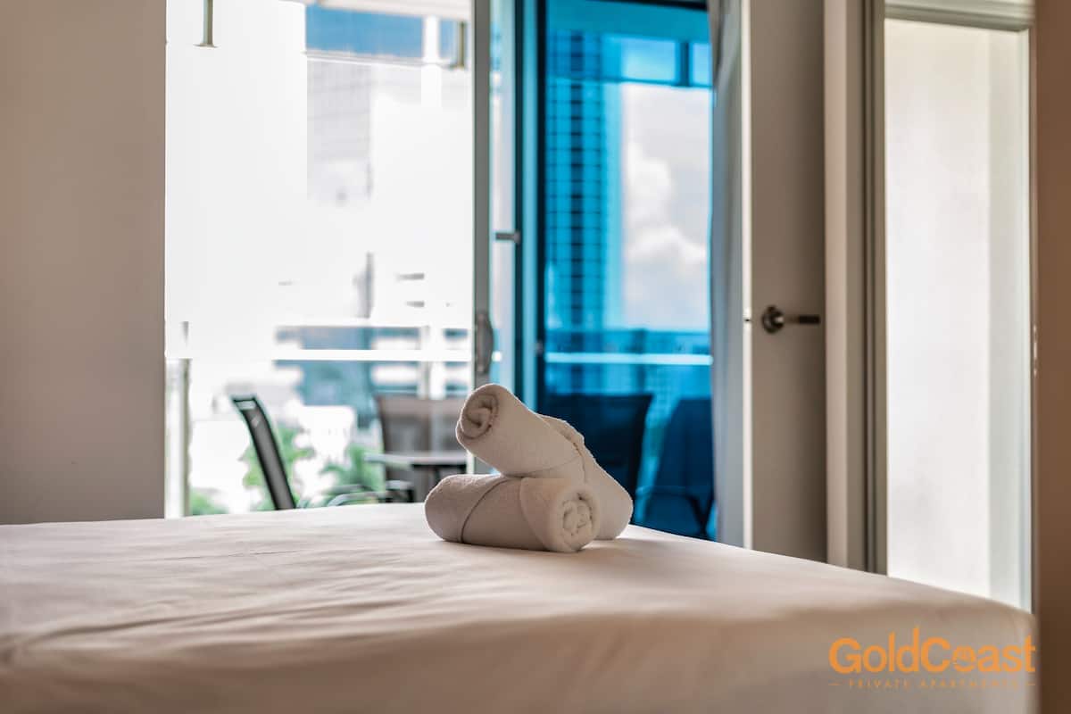 A neatly made bed with rolled towels in a hotel room with a view of a building through an open door.