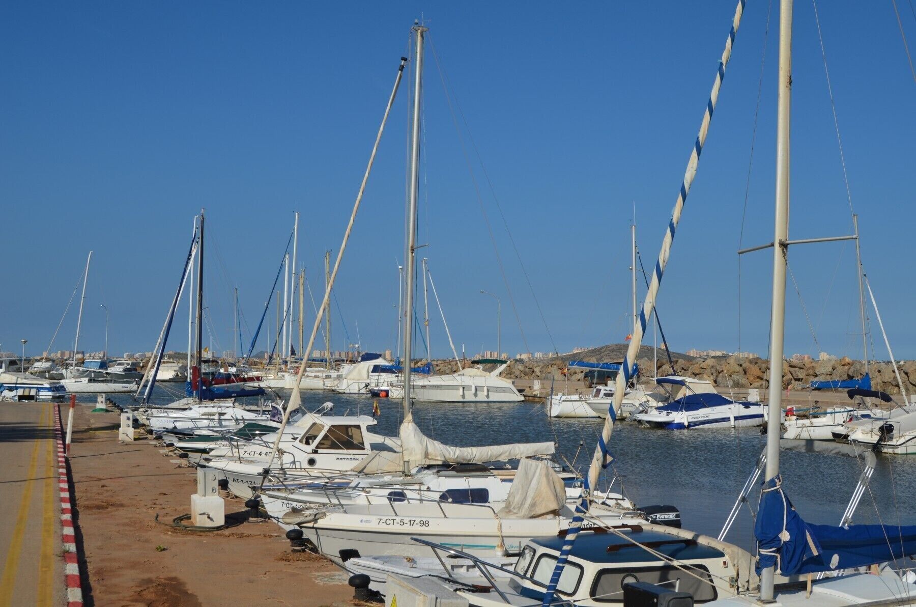 A marina with multiple sailboats docked, a sandy shore, and a clear blue sky.