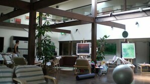 A spacious indoor area with high ceilings, wooden beams, and multiple seating areas including chairs and a sofa. There are several potted plants and a large exercise ball. - Gallery Court OFA ATU (Kawazu)
