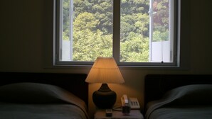 A bedroom with two beds, a nightstand with a lamp, a phone, and a view of a forest through a window. - Gallery Court OFA ATU (Kawazu)